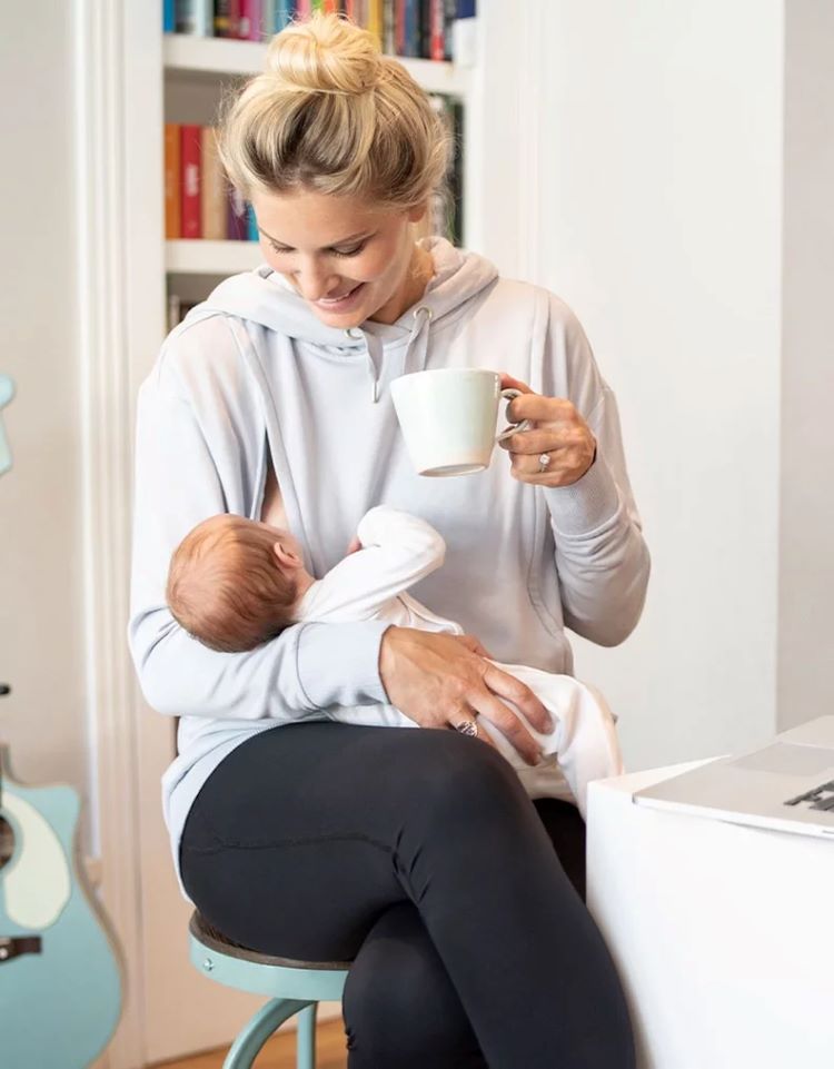 woman wearing maternity top sitting on a chair while drinking coffee and breastfeeding her baby 